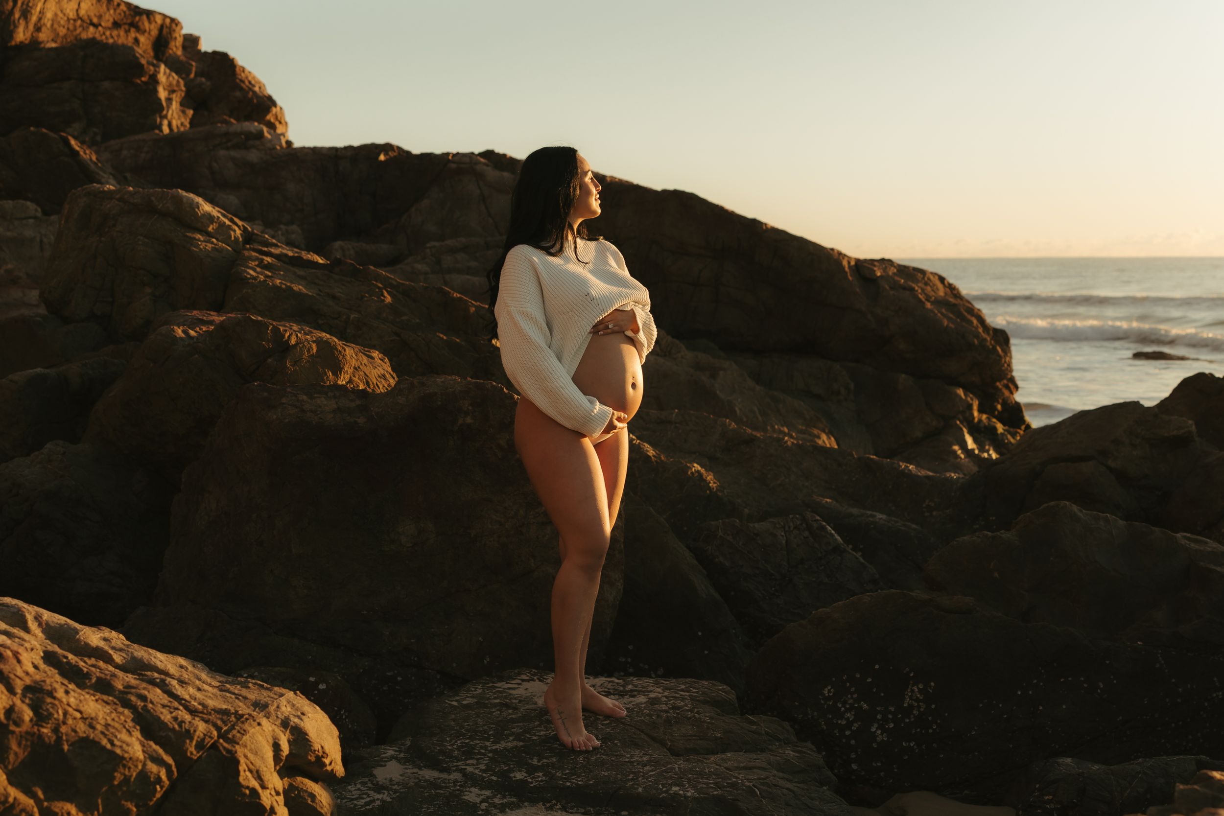 Couple standing on rocky coastal cliffs at sunset with ocean waves crashing below.