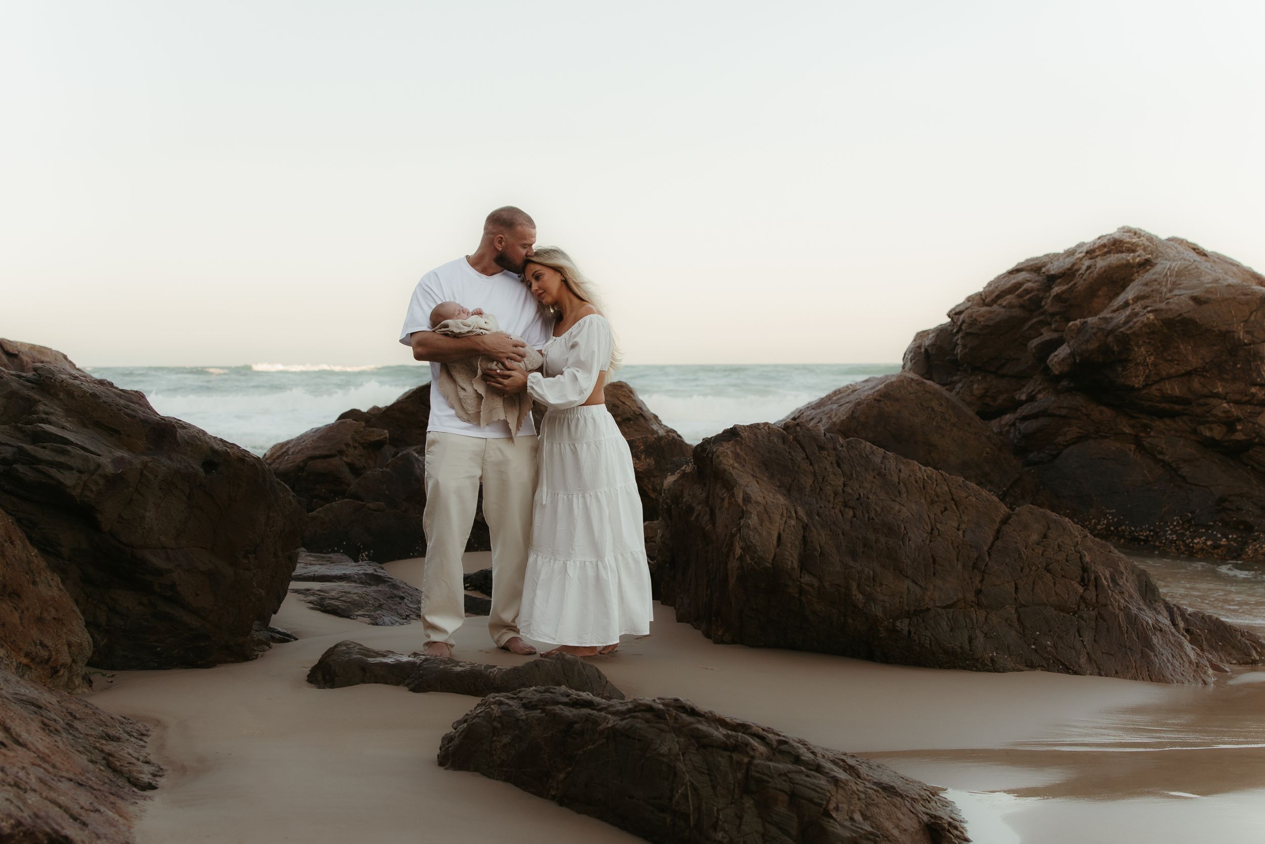 A romantic couple in white attire embraces on a rocky beach at sunset, creating silhouettes against the ocean backdrop.
