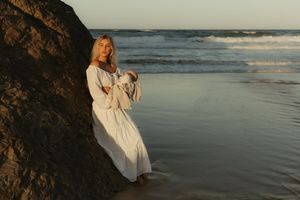 A person in a flowing white dress walks along a misty beach shoreline at sunset.