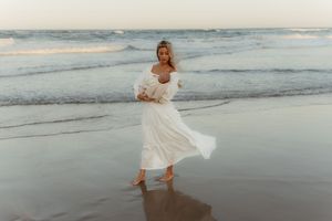 Someone in a flowing white dress twirls on a beach at sunset with ocean waves in the background.