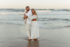 Two people in white outfits walking together on a beach at sunset with ocean waves in the background.