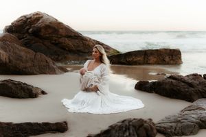 A person in a flowing white dress sits gracefully among rocky coastal formations as waves crash in the background.
