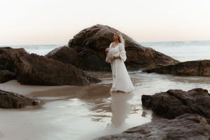 A figure in a flowing white dress walks gracefully along a rocky beach shoreline at sunset.