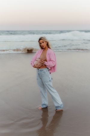Person in a pink jacket and light denim jeans stands on a beach at sunset with waves in the background.