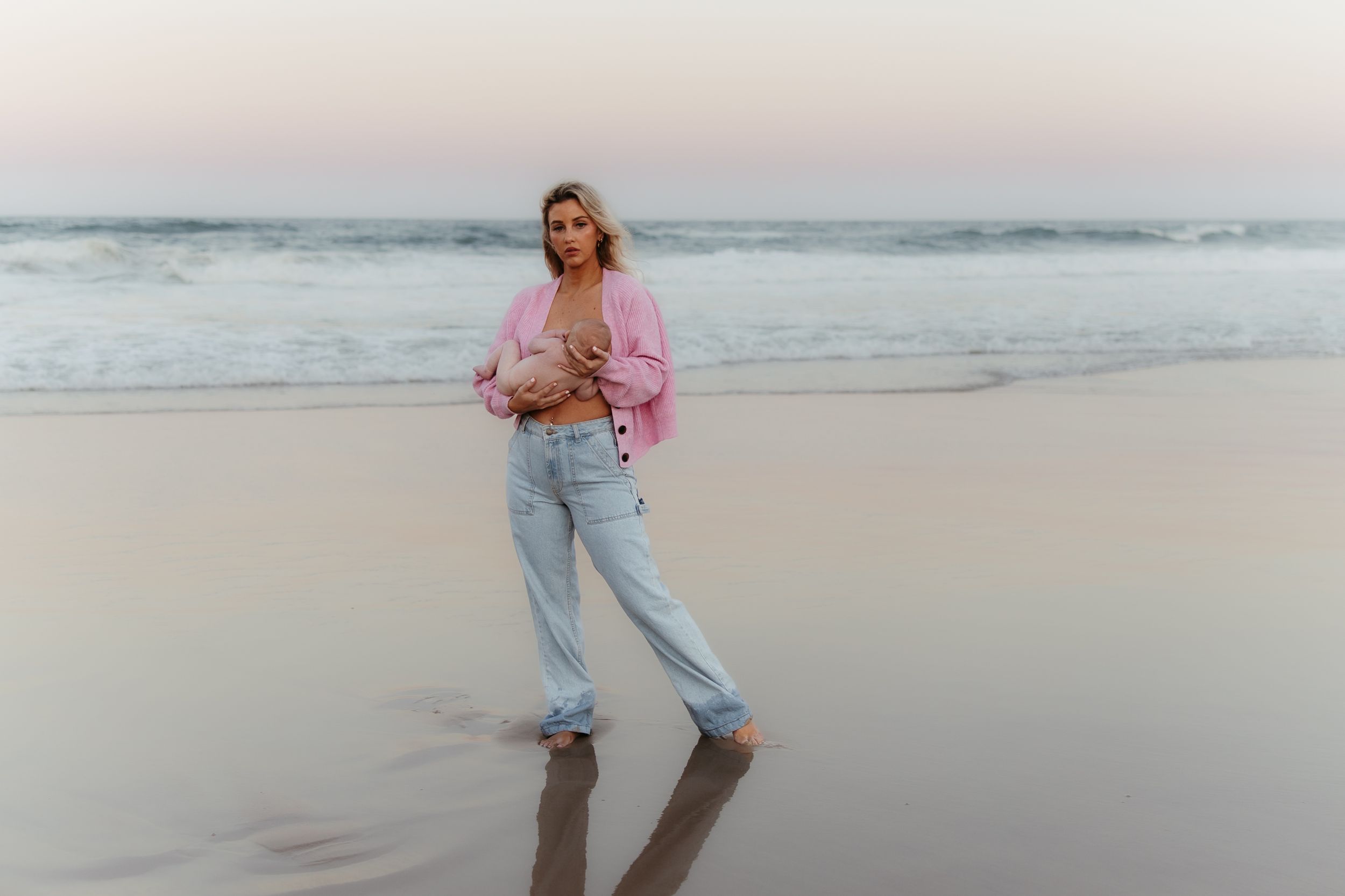 A person in a pink sweater and light jeans stands casually on a misty beach at sunset.
