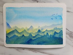 Watercolor mountain landscape painting with layered blue-green peaks and birds flying in a bright sky.