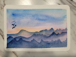 Misty mountain range watercolor painting in shades of blue with birds against a sunset sky.