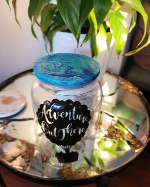 Glass jar with blue resin lid and adventure-themed text decoration next to a houseplant.