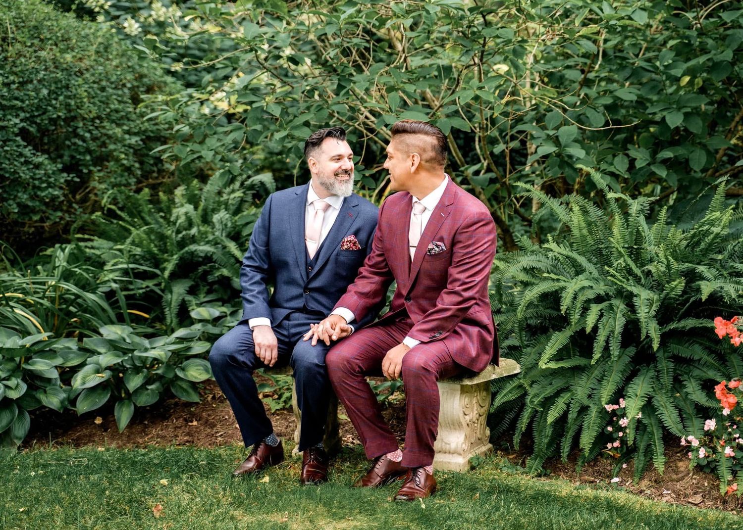 Two people in formal suits sitting on a stone bench in a lush garden setting with ferns and greenery.