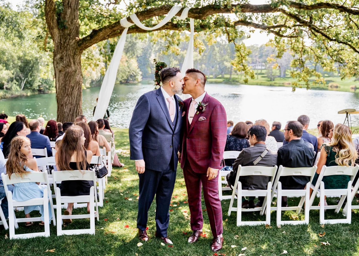 Outdoor wedding ceremony under a grand tree with white drapes and folding chairs set up on a lawn by a lake.