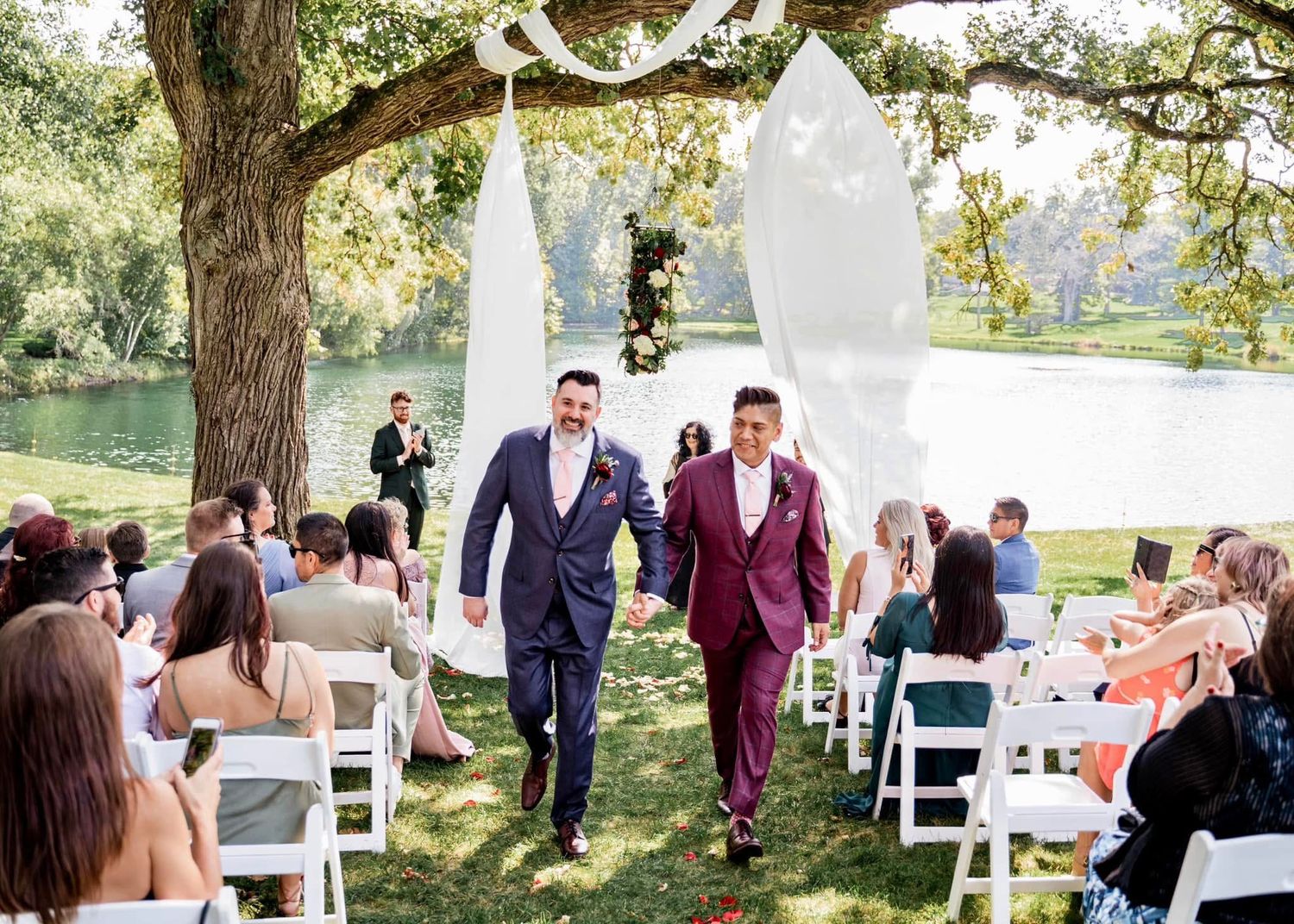 Two grooms walk hand in hand down the aisle at an outdoor wedding ceremony beside a lake with white chairs and decor.
