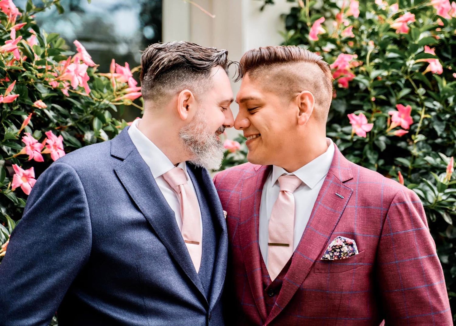 Two people in formal attire share an intimate moment in front of pink rose bushes.