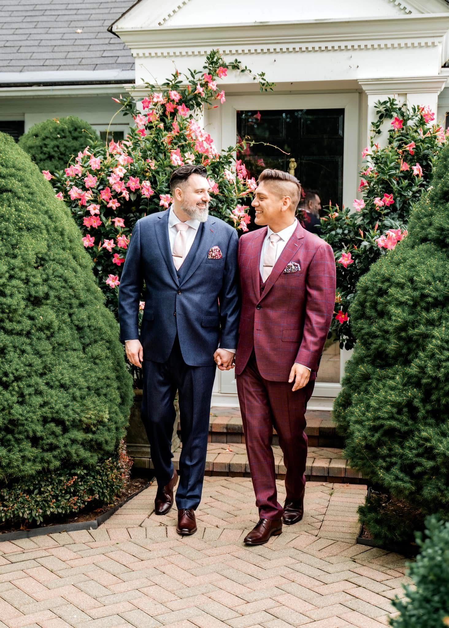 Two people in formal suits walk together on a brick path framed by pink rose bushes and manicured shrubs.