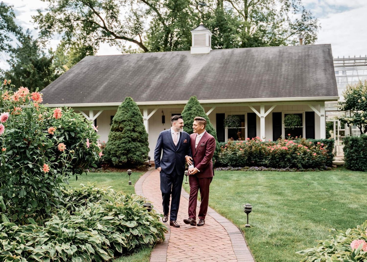 Two people in elegant formal attire walk along a brick path towards a charming cottage-style home with landscaped gardens.