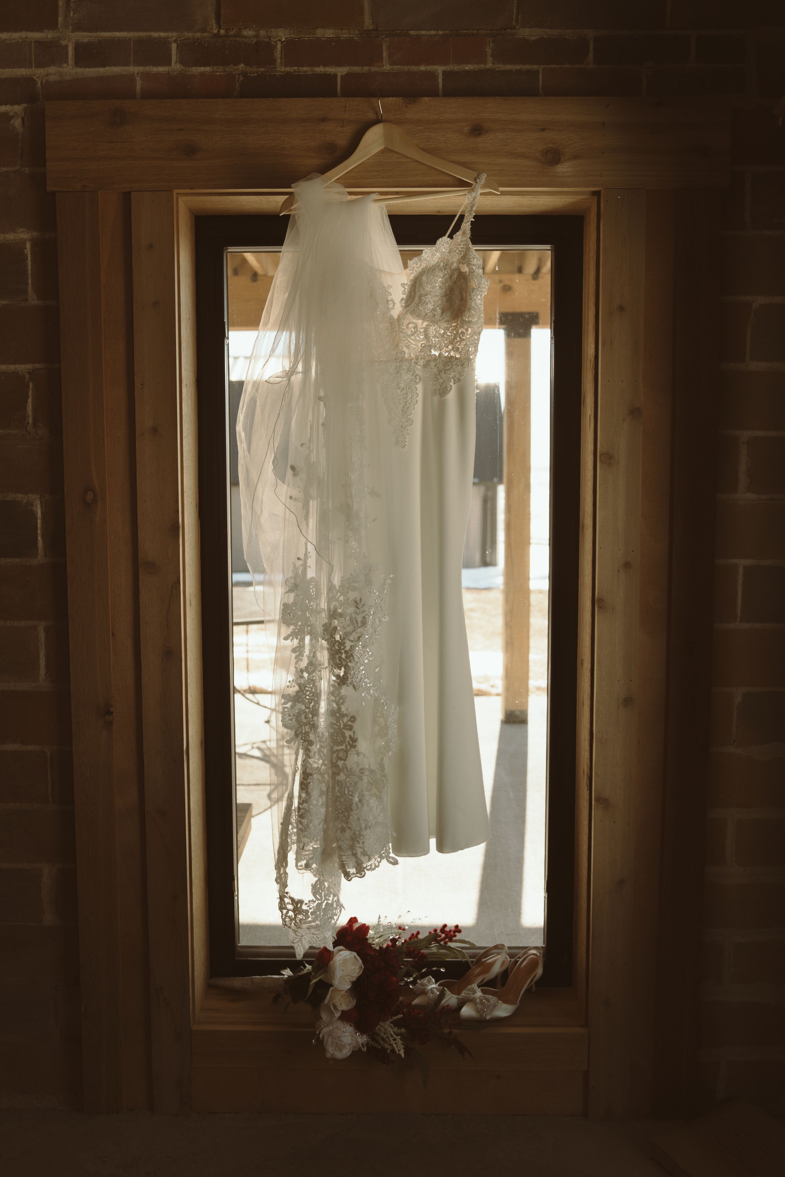 A white wedding dress hangs in front of a window with brick walls creating a silhouette.