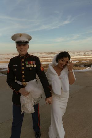 A couple poses together on a beach, with one wearing a Marine dress uniform and the other in a white wedding dress.