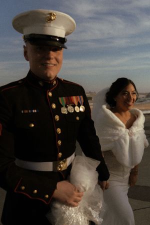 A couple embraces on a beach at sunset, with one in Marine dress blues and the other in a white wedding gown.
