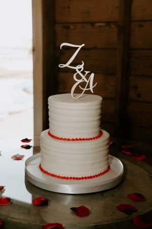 White wedding cake with red trim and decorative monogram topper displayed on wooden surface.