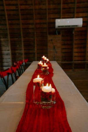 Long table decorated with red table runner and lit candles for wedding reception.