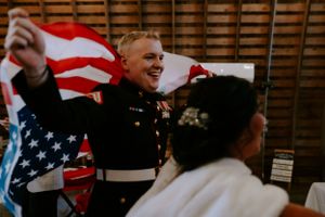 A military service member celebrates at a wedding reception with an American flag in the background.