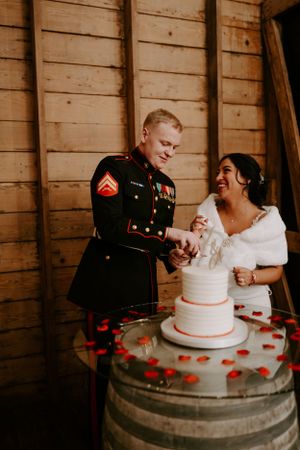 A military couple cuts their wedding cake together on a rustic wooden barrel.