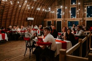 Wedding reception guests seated at tables with red decor inside a rustic barn venue.