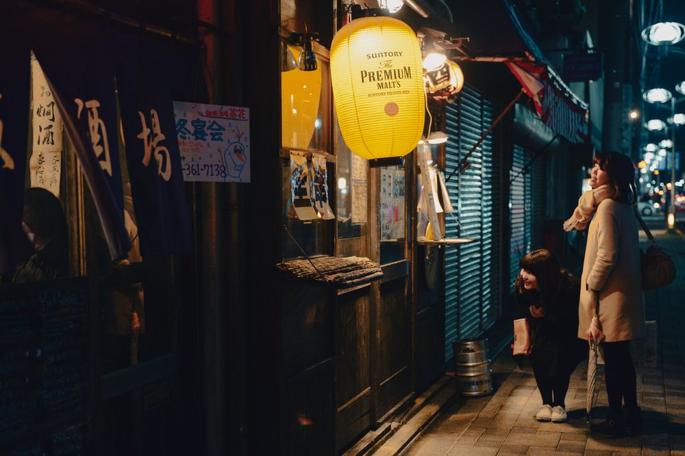 Nighttime street scene in Japan with illuminated shop signs and pedestrians walking.
