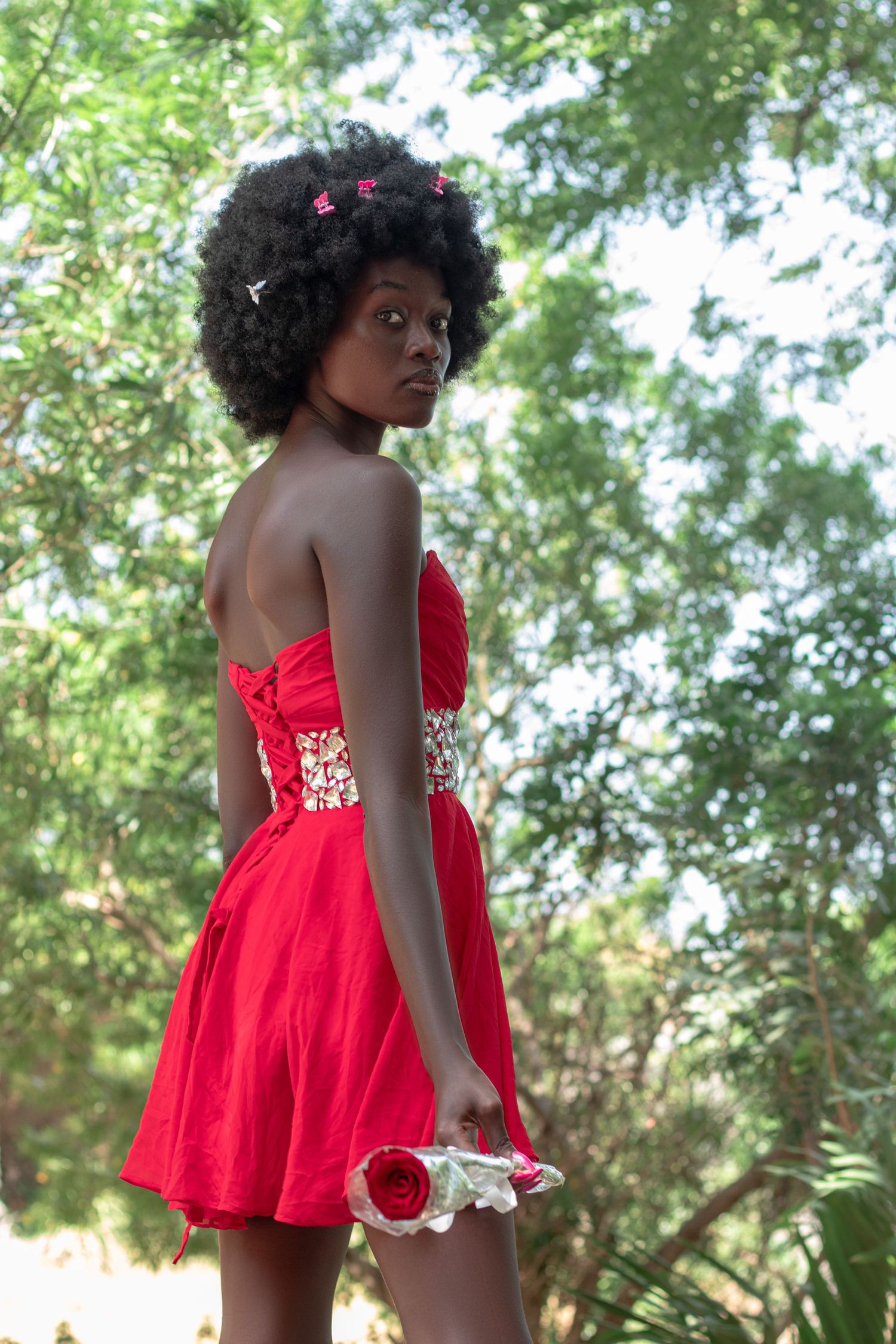 Lady poses with flower at her hand her body facing away from camera her face slightly bent torwards the camera.