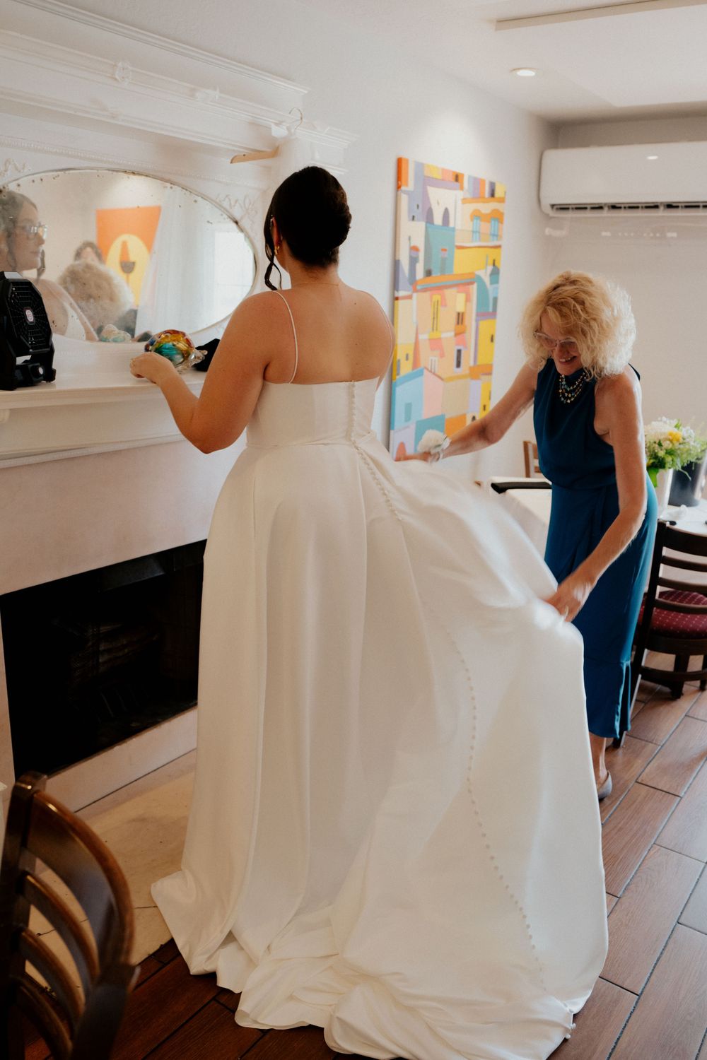 Someone in an elegant white wedding gown gets help with final dress adjustments in a bright room with colorful artwork.
