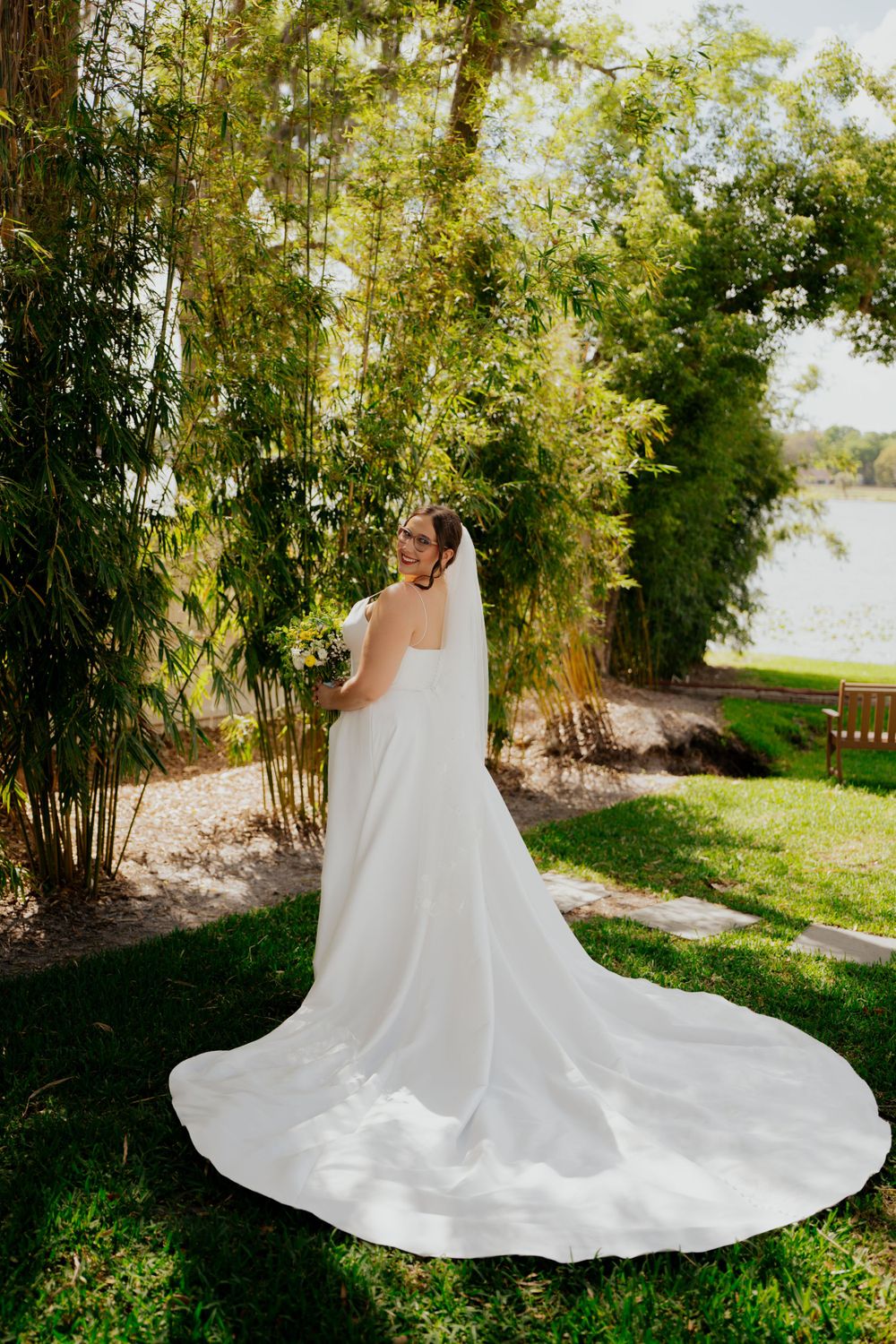 A stunning white wedding dress with a long flowing train photographed in a garden setting with bamboo and sunlit trees.