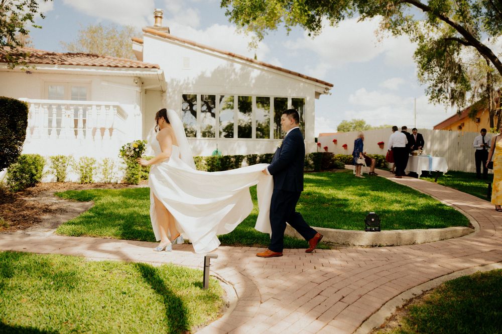 A couple in wedding attire holding a flowing white dress train on a garden path in front of a Spanish style villa.