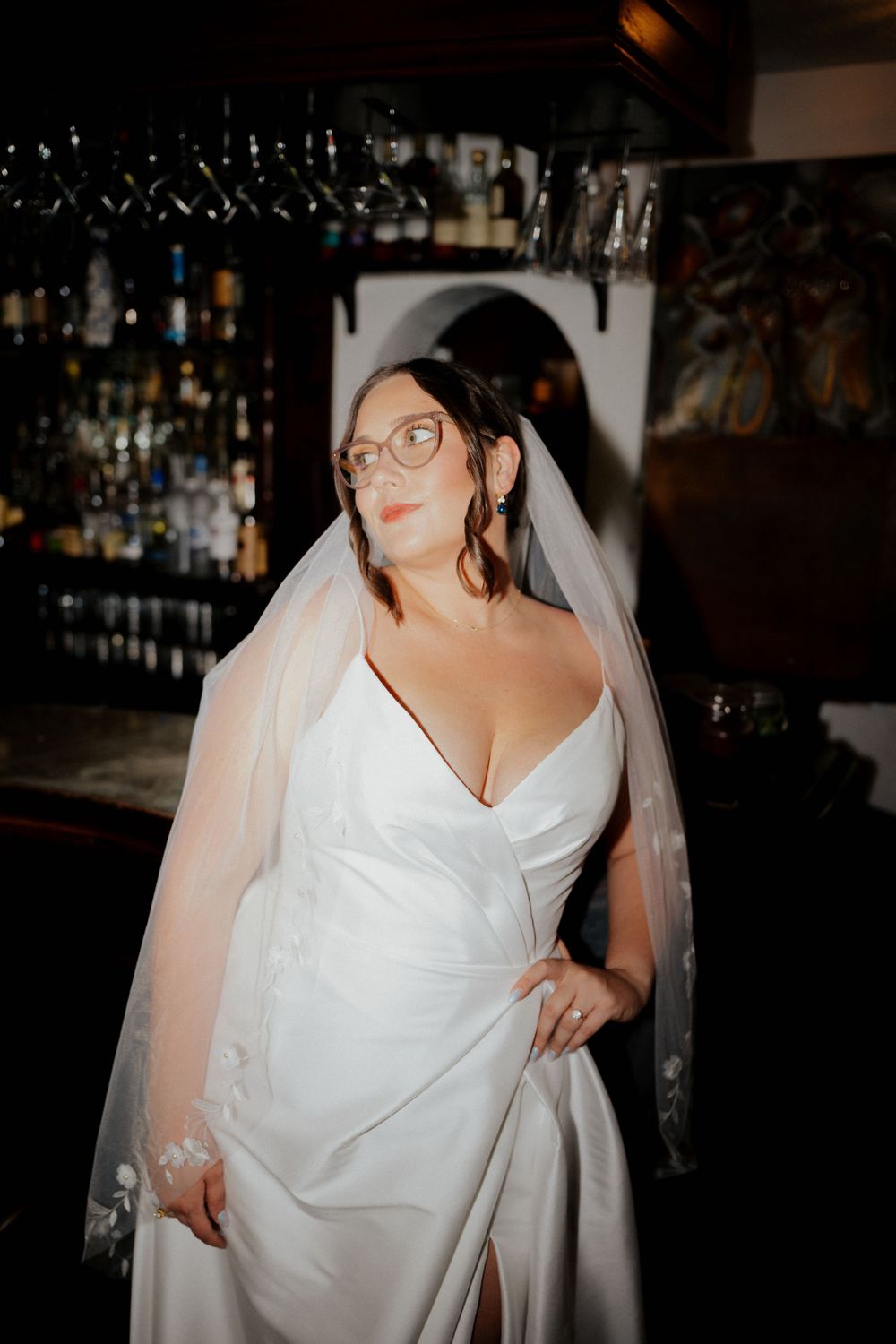 A bride in a classic white wedding gown and veil poses at a enzos on the lake bar with glasses and bottles in the background.