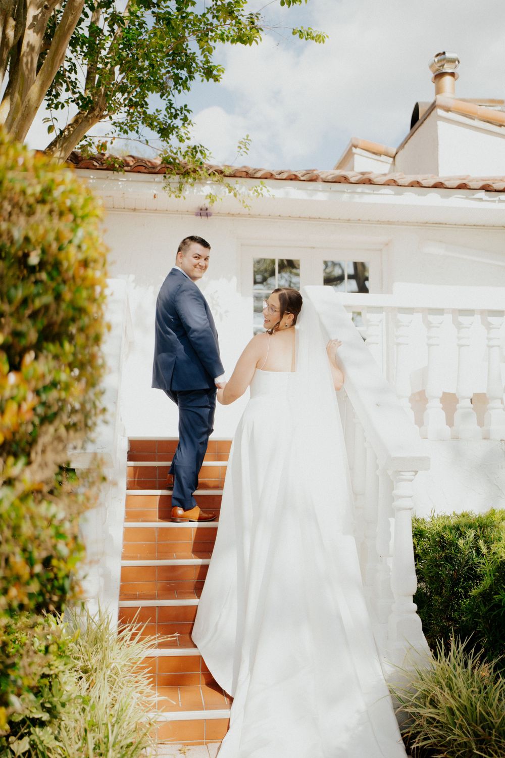 A wedding couple stands on terracotta stairs against a white Mediterranean-style building surrounded by lush greenery.