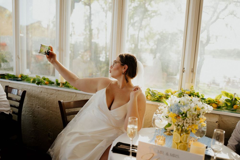 A bride in a white gown takes a selfie while seated at a table decorated with yellow flower arrangements.