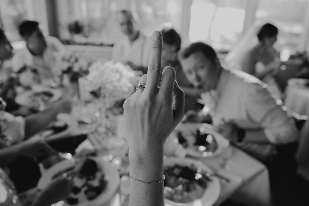 A raised middle finger gestures in black and white at a busy dining table scene with people eating in the background.