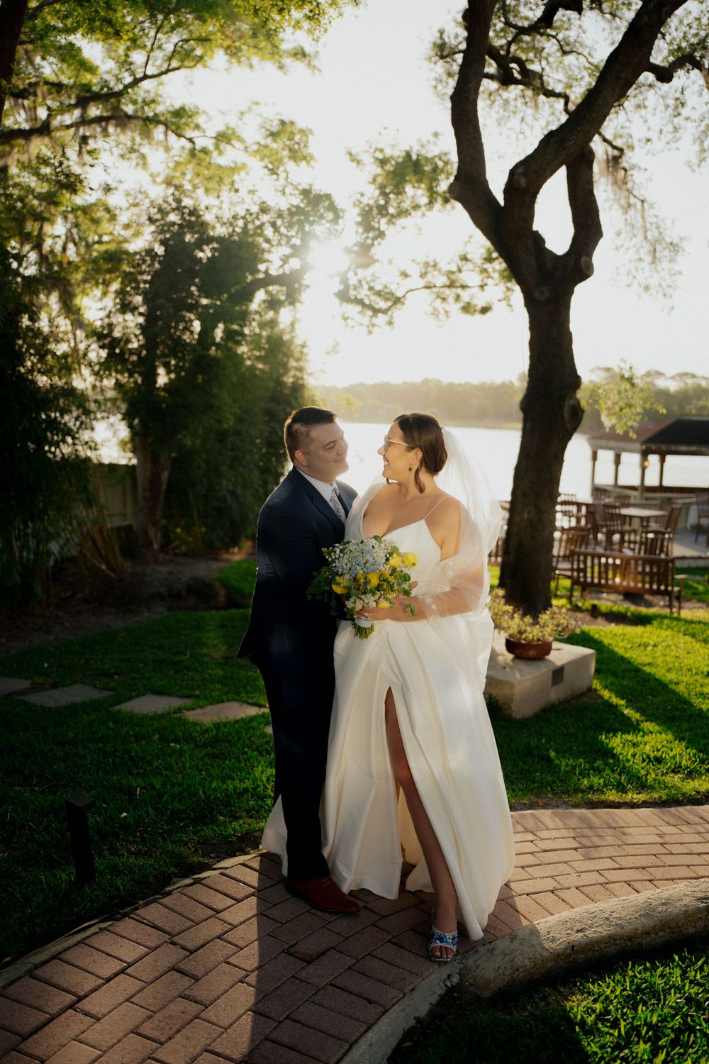Couple shares romantic moment at sunset on brick pathway near waterfront with tall tree and garden setting.
