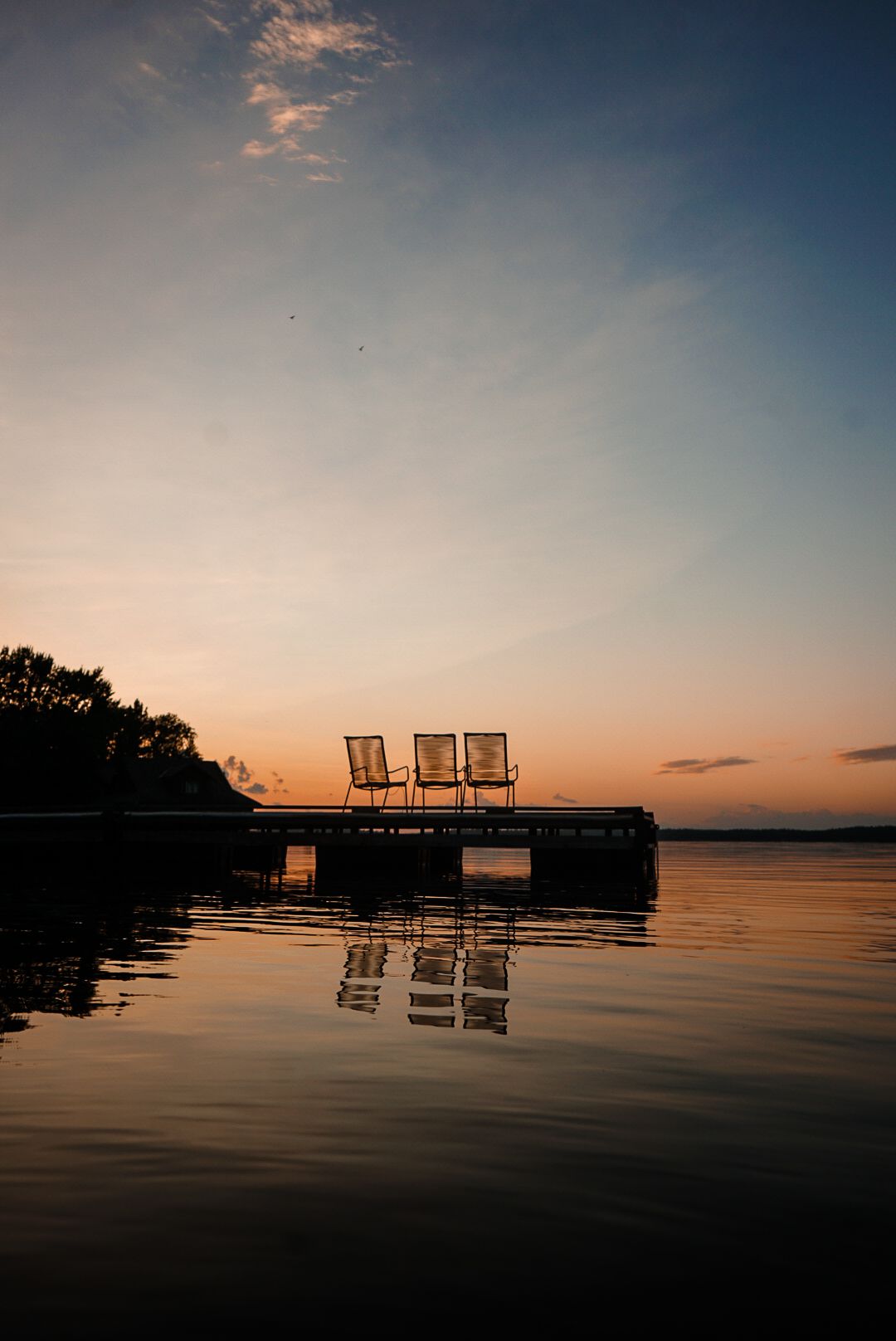 Silhouettes of chairs reflected in calm lake waters during sunset.