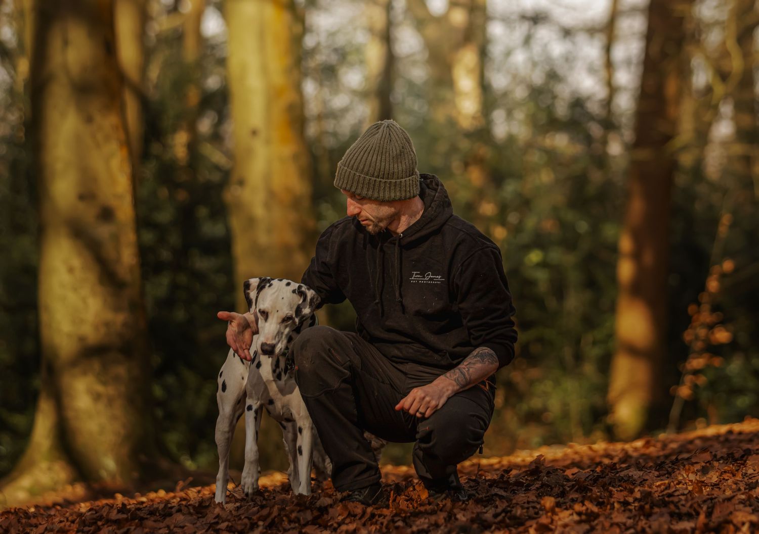 A person in a beanie and dark jacket crouches next to a spotted Dalmatian dog in an autumn forest.