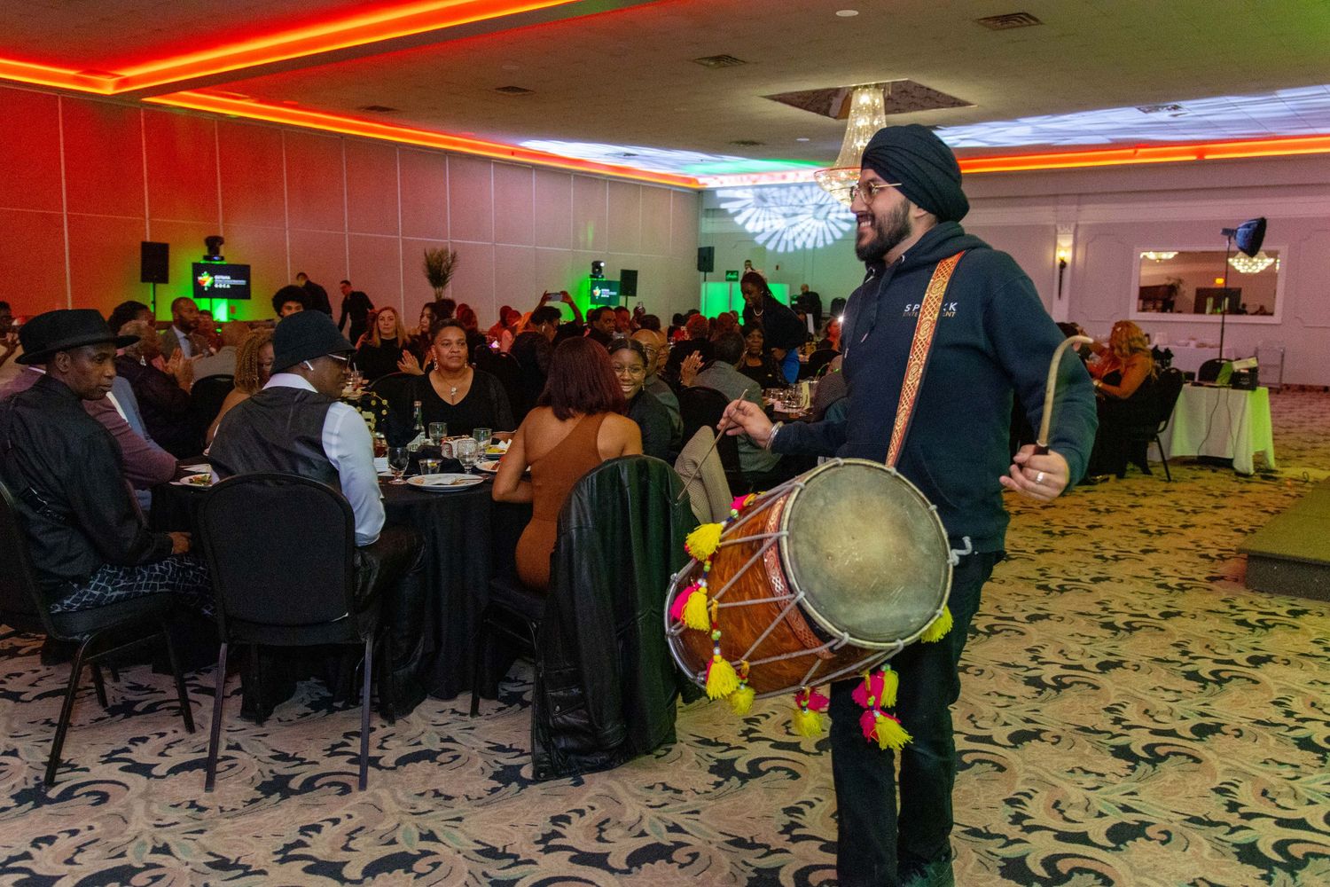A musician holding a traditional drum performs at a festive banquet hall event with colorful lighting and seated guests.
