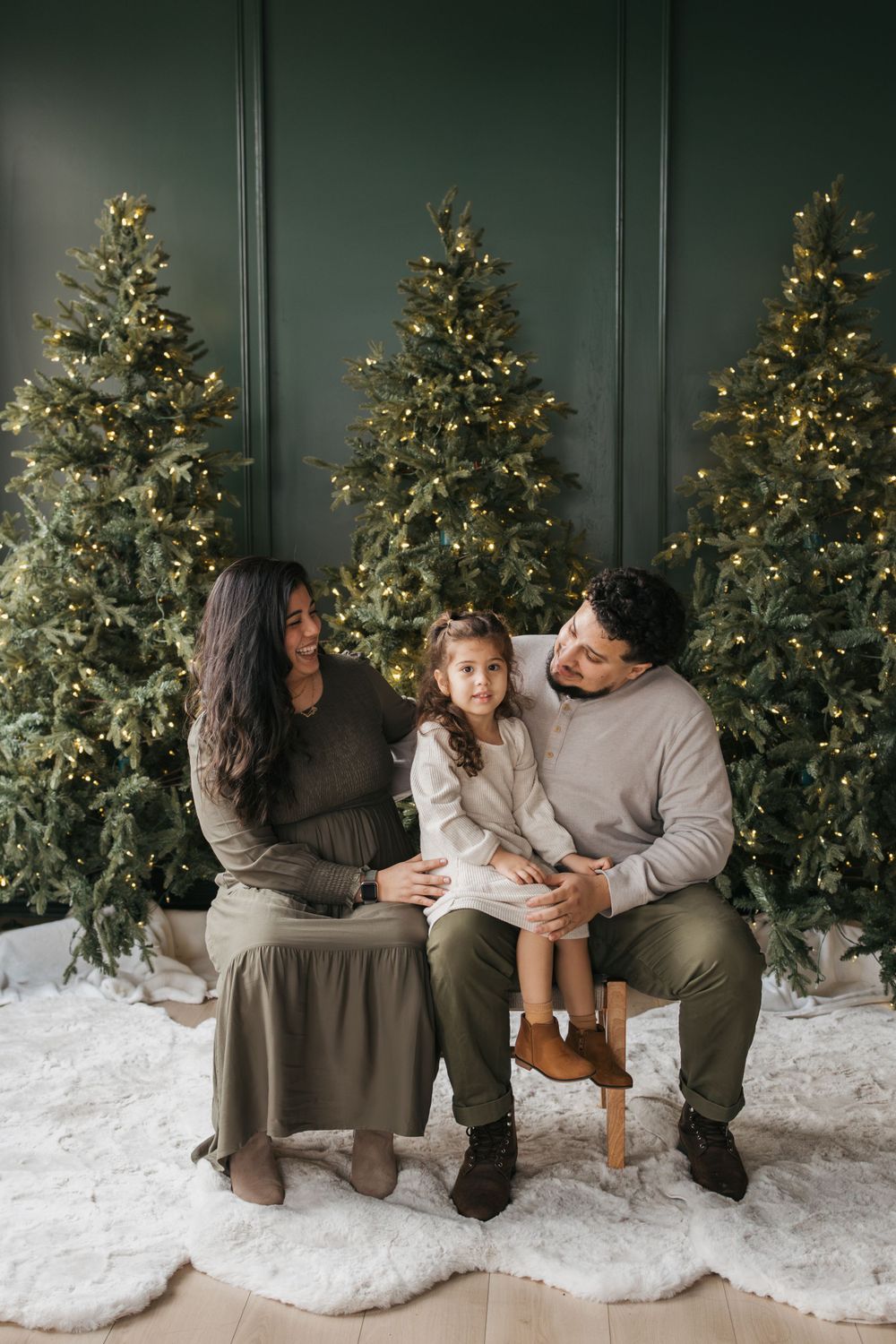 A family sits together in front of three illuminated Christmas trees on a white faux fur rug for a holiday photo.