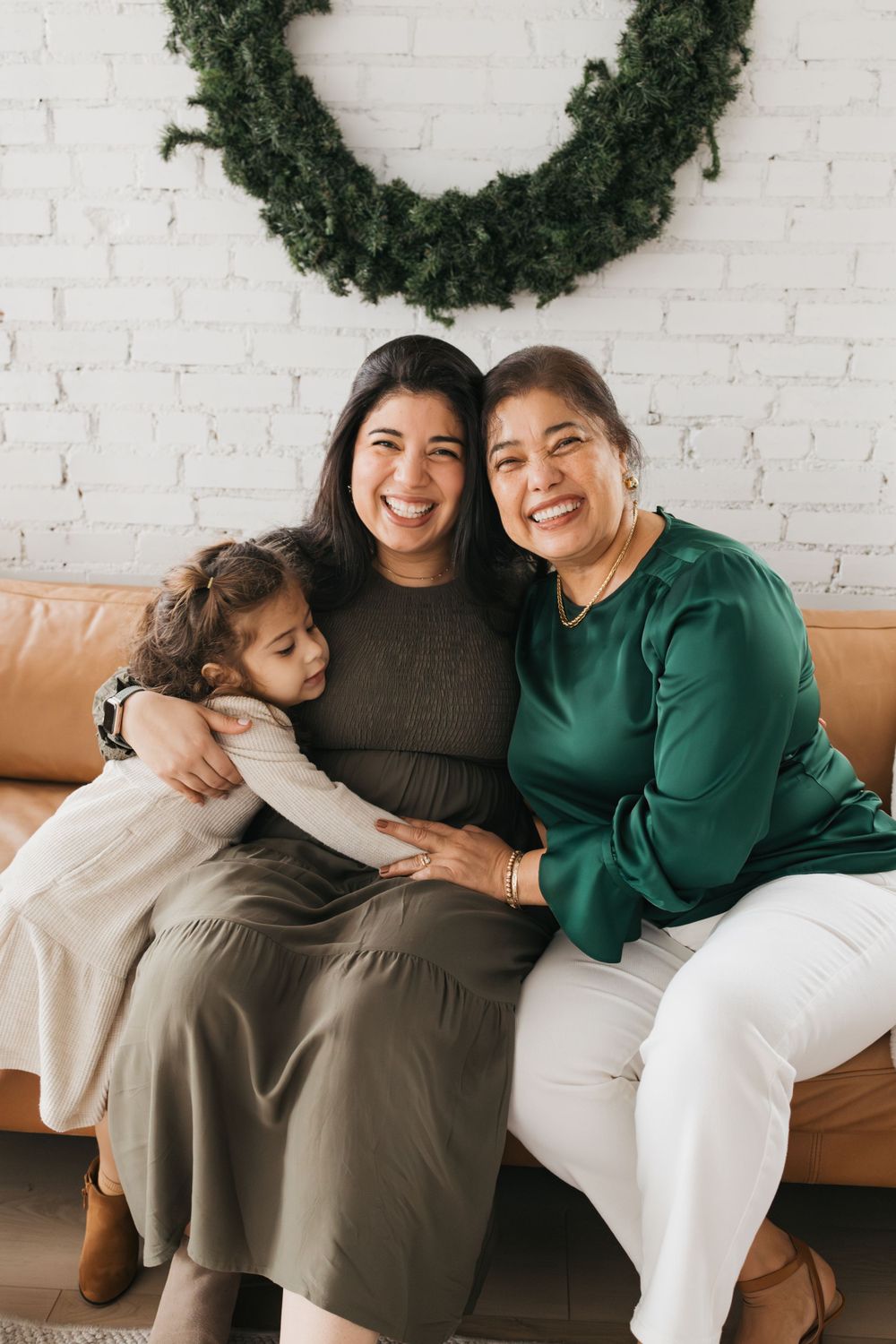 Three generations share a joyful moment on a tan leather couch under a festive wreath on a white brick wall.