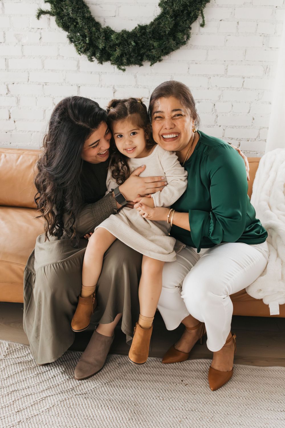 Three people sit together on a tan leather couch under a holiday wreath in a cozy home setting.