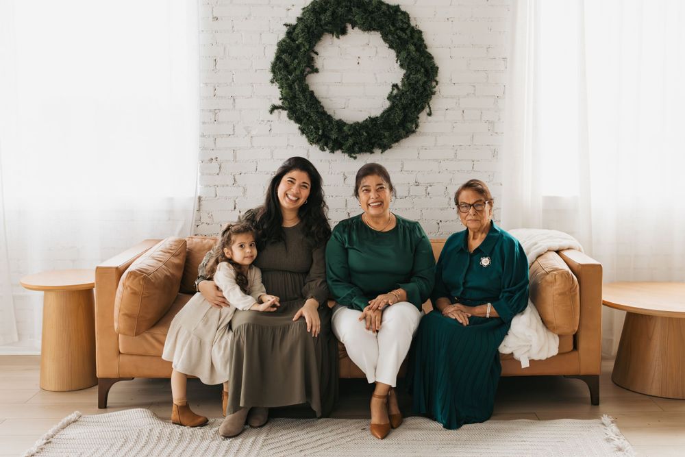 Three people sit on a tan leather couch under a festive green wreath on a white brick wall.