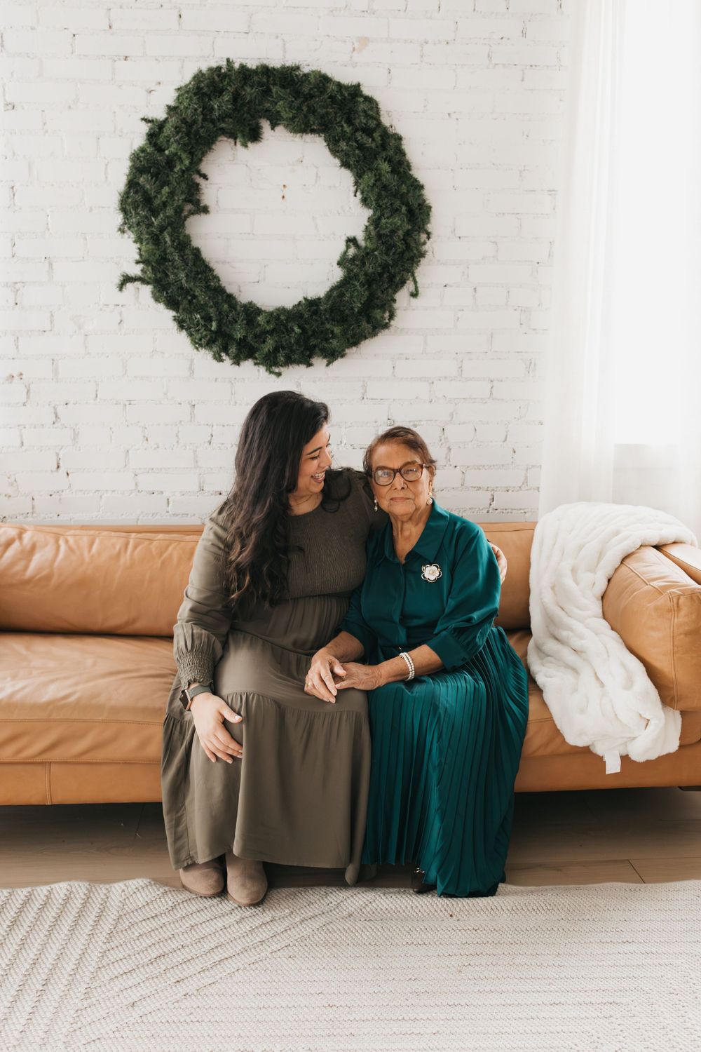 Two people sharing a tender moment on a tan leather couch beneath a green wreath on a white brick wall.