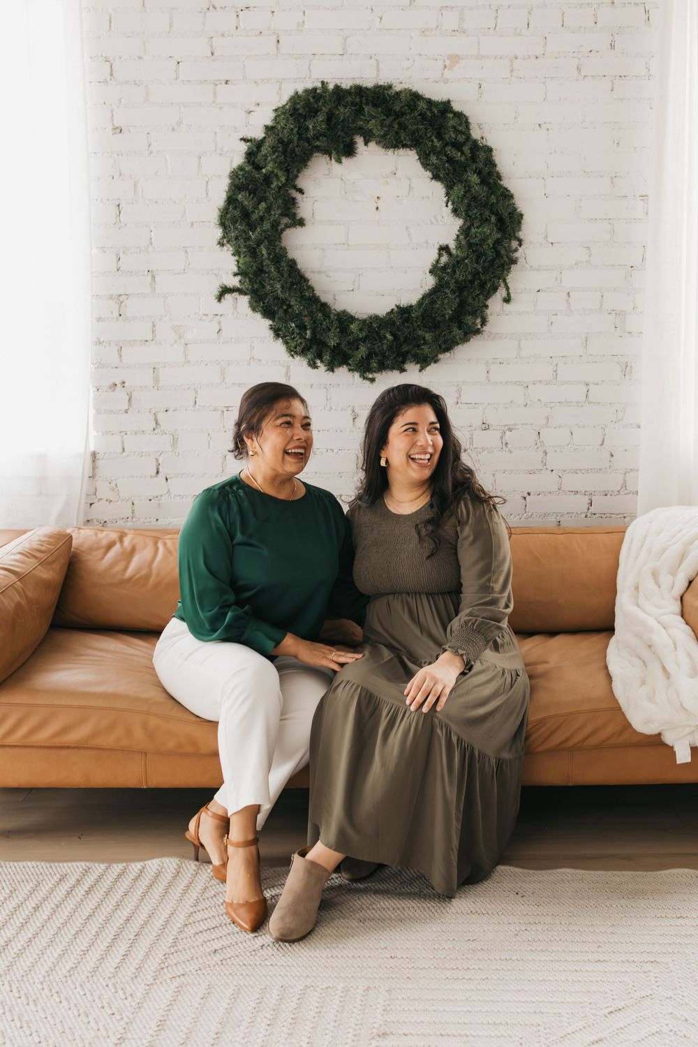 Two friends share a joyful moment on a tan leather couch beneath a minimalist evergreen wreath.
