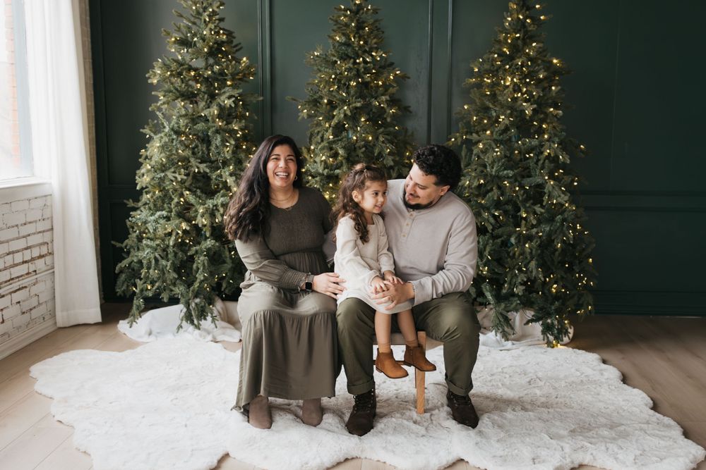 A family sits together on a white rug in front of three decorated Christmas trees in a cozy indoor setting.