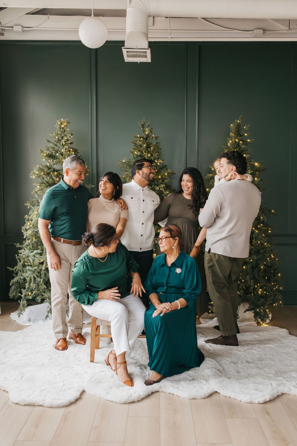 Multi-generational family gathering posed in front of three decorated Christmas trees against a green wall.