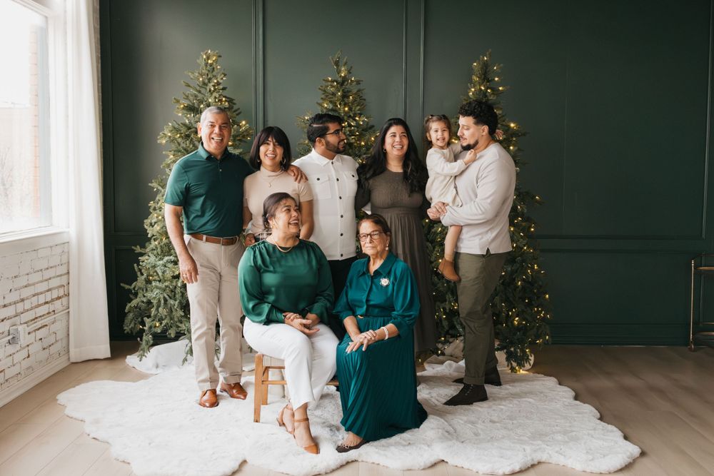 A group poses in coordinated green and white outfits between two lit Christmas trees against a dark green wall.