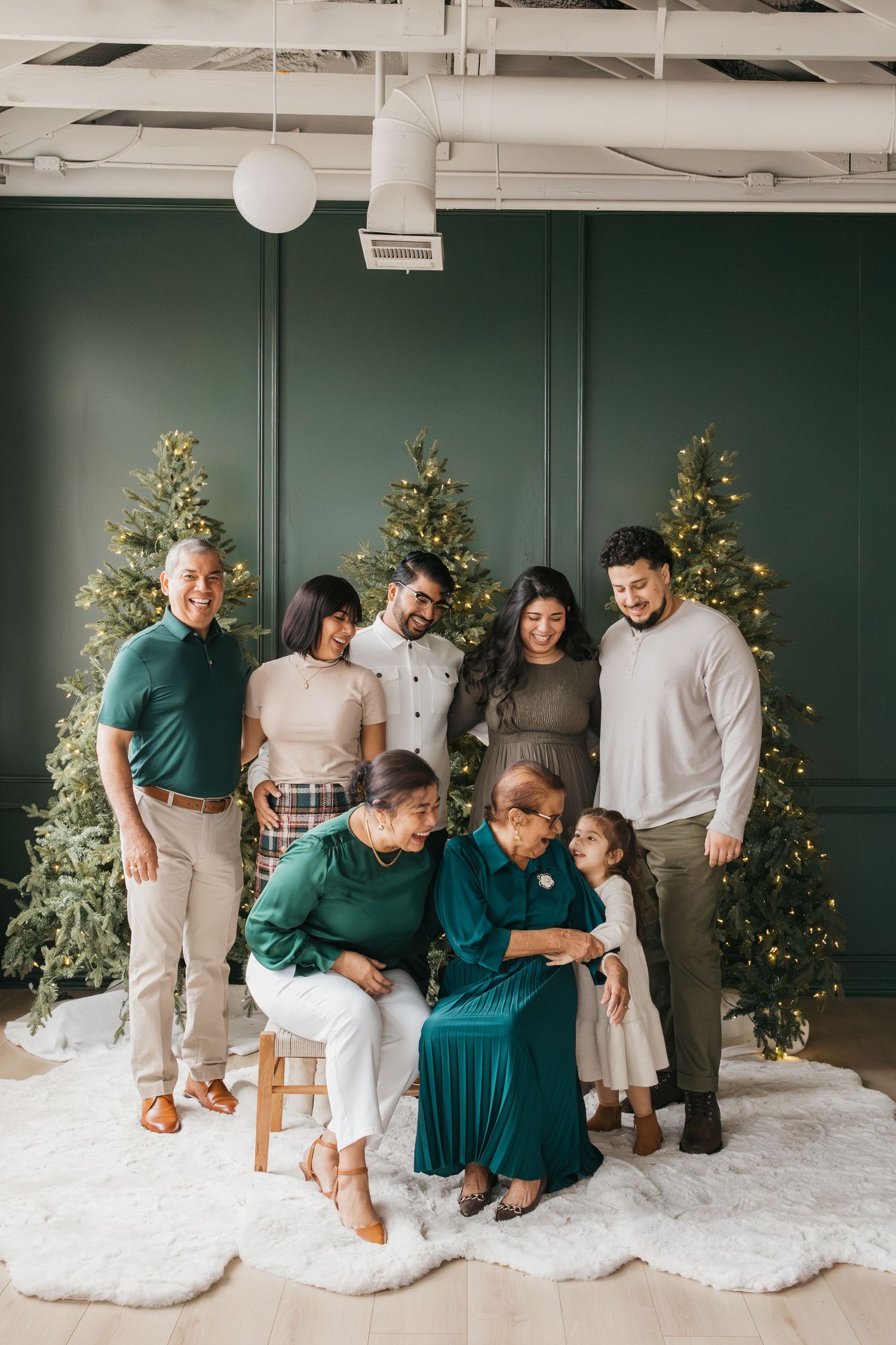 Family group poses in coordinated green and white outfits in front of three lit Christmas trees on white carpet.