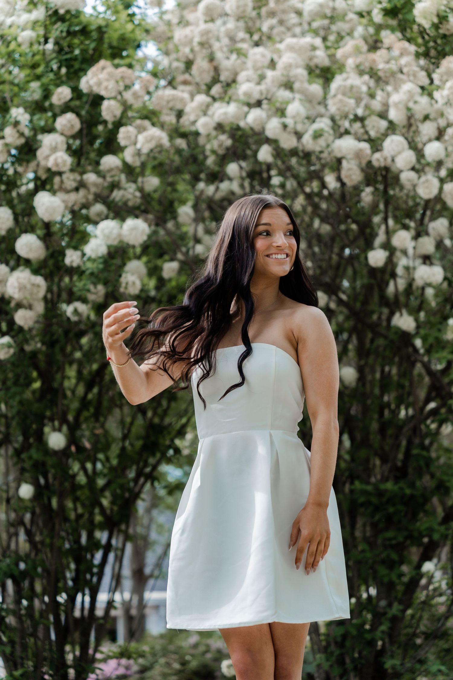 A woman in a white strapless sundress smiles joyfully while standing in front of blooming white floral bushes.